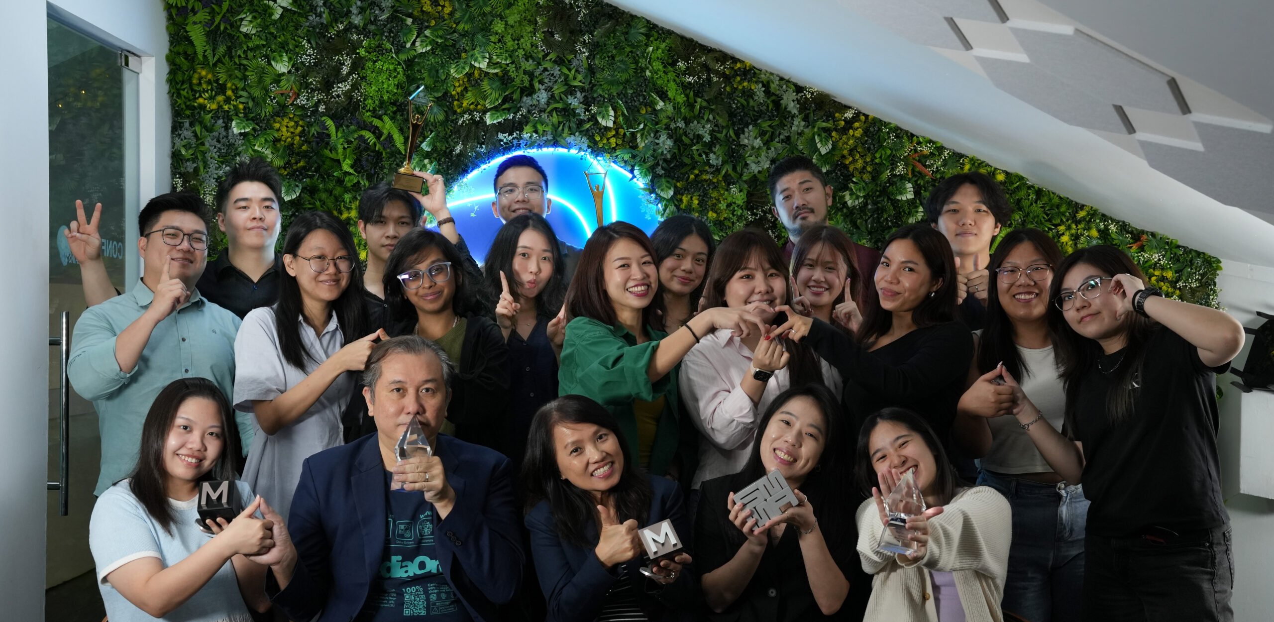 A large group of diverse Asian professionals, men and women, celebrating with multiple awards and trophies against a vibrant green plant wall and blue neon background of MediaOne Company
