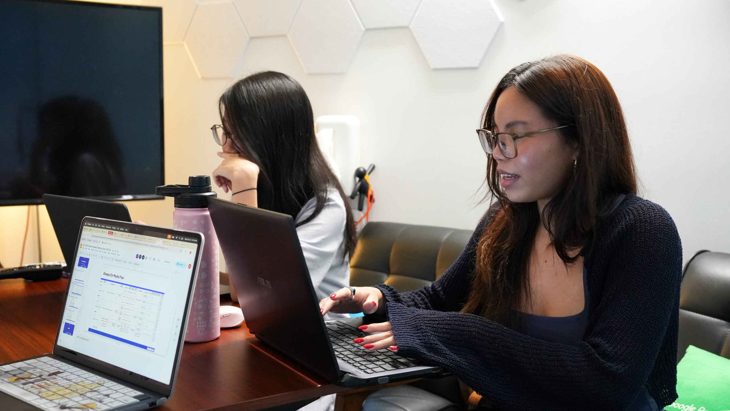Two women focused on their laptops at a shared desk, one typing and the other contemplating her screen, a document titled 'Always On Media Plan' visible on a laptop.
