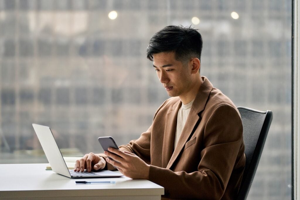 Young Asian man multi-tasking with laptop and smartphone in modern office